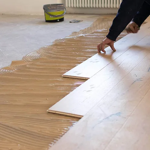 Worker applying adhesive and laying down wooden flooring planks in a well-lit interior space.