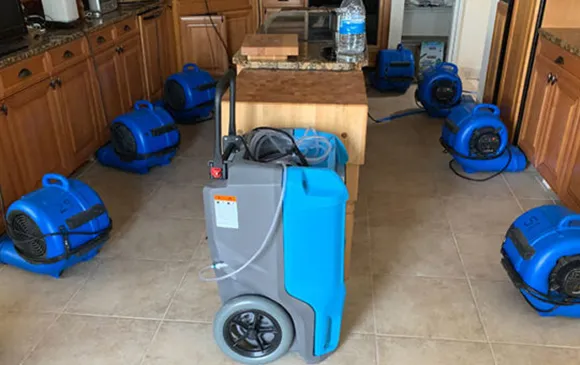 Water damage restoration equipment set up in a room, including multiple blue air movers and dehumidifiers.