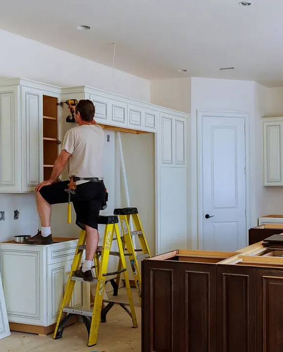 Woman standing on a ladder while installing cabinetry and molding in a kitchen remodel.