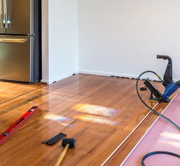 Worker laying down wood flooring planks with tools in a bright room.